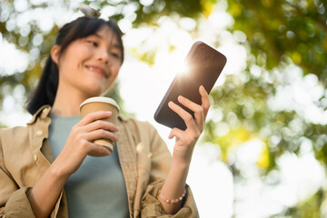 Happy young female freelancer holding paper cup and using smartphone in the summer park