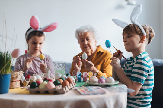 Grandmother with little kids decorating easter eggs at home. Tradition of painting eggs with brush and easter egg dye.