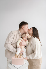 Man kiss woman. Father, mother hugs funny little kid. Family celebrates six month old baby girl birthday party closeup. Dad and Mom hold hands cute baby isolated on white wall. Creative birthday cake.
