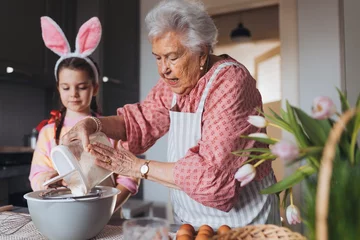 Fototapeten Grandmother with granddaughter preparing traditional easter meals, baking cakes and sweets. Passing down family recipes, custom and stories. © Halfpoint