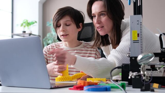 Happy Caucasian mother helping son with homework, doing school project together, using laptop, 3D printer and printed details of prototype, sitting at desk on table.