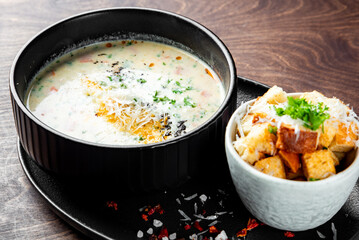 A creamy soup with herbs and spices, served in a black bowl, accompanied by croutons in a small bowl on a wooden surface. The background is a dark wood