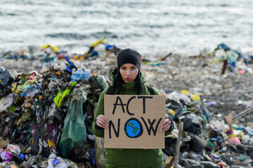 Activist holding placard, protest sign, standing on landfill, large pile of waste on sea shoreline,...