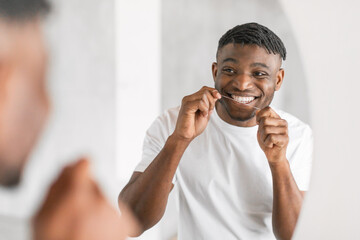 Smiling young black man flossing teeth near mirror in bathroom