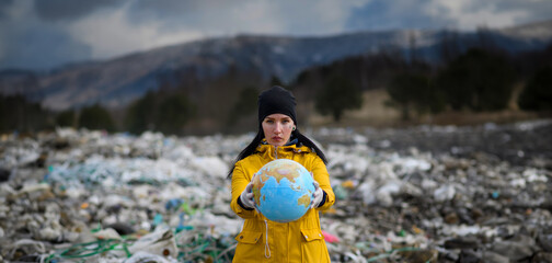 Activist holding Eart model, globe, standing on landfill, large pile of waste on sea shoreline,...
