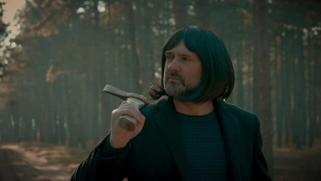 Man with axe on his shoulder walks through forest disguised as killer with wig to remain unrecognized. Maniac Murder and crime, Halloween horror day concept