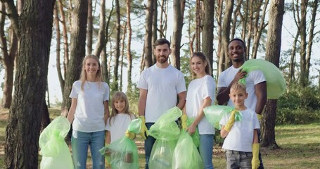 Attractive multiethnic people man woman and children looking at camera after cleaning park from scattering rubbish in plastic bags. Protection of environment concept