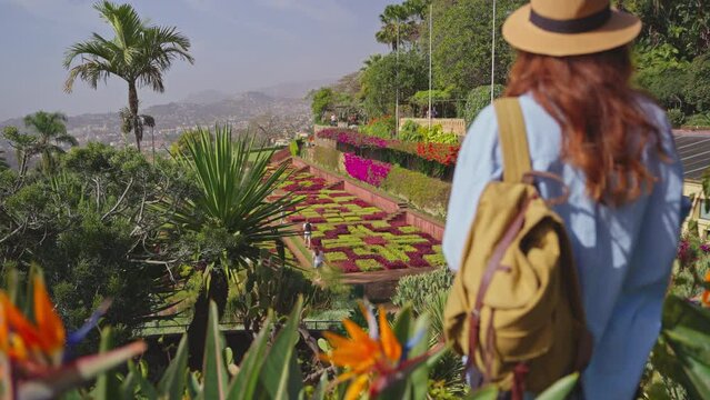 Traveler visiting a landmark garden with diverse vegetation of the island Madeira and Funchal city. Woman with hat and backpack walking between colorful flowers in botanical garden