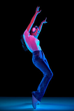 Full-length image of young girl in casual clothes, white t-shit and jeans listening to music in headphones and dancing against black background in neon. Concept of youth, lifestyle, fashion, emotions