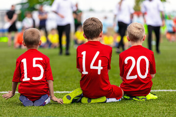 Three school boys sitting on the pitch line wearing red soccer jerseys with white numbers. Happy kids play football tournament game
