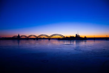 Zelfklevend Fotobehang A beautiful sunrise scenery with iron bridge over the frozen river Daugava in Latvian capital city Riga. Winter landscape of Northern Europe. © dachux21