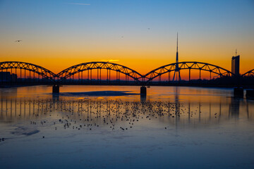 Fototapeta na wymiar A beautiful sunrise scenery with iron bridge over the frozen river Daugava in Latvian capital city Riga. Winter landscape of Northern Europe.