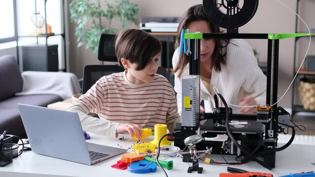 Curious Caucasian son and mother control work of a 3D printer together at home workshop. Education and collaboration in family, parent and child teamwork concept.