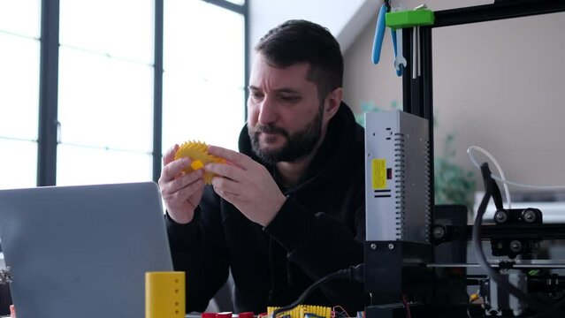 Serious male engineer assembly and testing cogwheel mechanism, during work with laptop and 3D printer in home office. Analyzing future project.