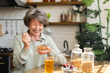 Cheerful senior grandmother eating cereals for breakfast mixing with berries drinking apple juice...