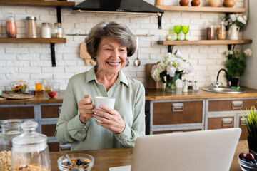 Smiling happy caucasian old elderly woman having healthy breakfast at home kitchen while watching...