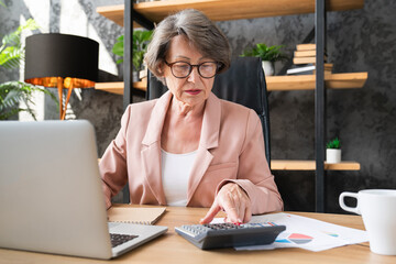 Concentrated caucasian senior businesswoman manager counting funds earnings bills on calculator in office. Serious grandmother paying domestic bills, loan, mortgage, insurance