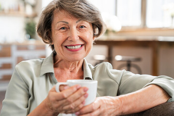 Cheerful caucasian old senior grandmother drinking tea coffee looking at camera. Elderly woman...