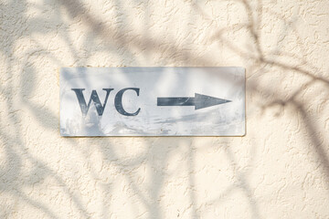 Faded WC sign pointing right, mounted on a textured wall with shadows of tree branches, indicating a restroom location