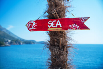 Hand-painted arrow sign with floral patterns indicating the direction to the sea, attached to a palm tree on a bright sunny day