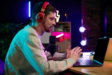 Young man sitting in home office in headphones, with microphone and laptop, recording online...
