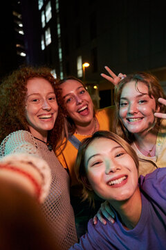 Four happy smiling young female friends taking a vertical selfie looking at camera together at night outdoors. Diverse group of women posing to self photo having fun at street on city.