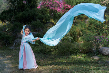 Fototapeta premium Pretty Asian girl in a beautiful elegant ancient Chinese costume traditional dress dancing with a flowing shawl made of thin long fabric pastel color waving in the wind at a green natural garden. 