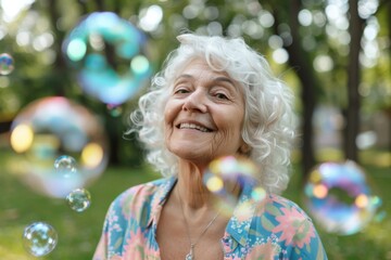 A senior woman with curly white hair and a colorful blouse smiles joyfully amidst floating soap bubbles in a lush park, evoking delight and nostalgia.