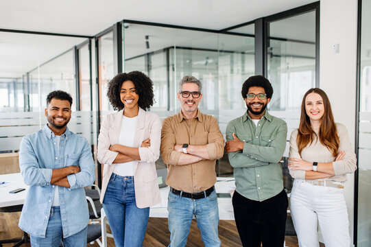 A vibrant team of five professionals stands with confidence in office, arms crossed and smiles wide, representing the epitome of a diverse and dynamic workgroup poised for collaboration and success