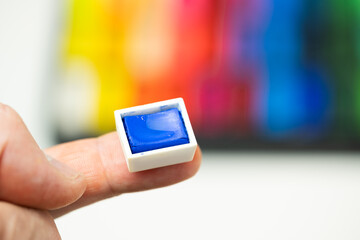 A finger showing a mini blue watercolor cube with a multicolored watercolor background. Landscape format.