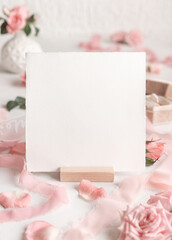 Blank square card near light pink roses, petals and silk ribbons close up, wedding mockup