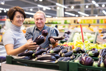 Couple standing in salesroom of greengrocer and choosing eggplant - obrazy, fototapety, plakaty