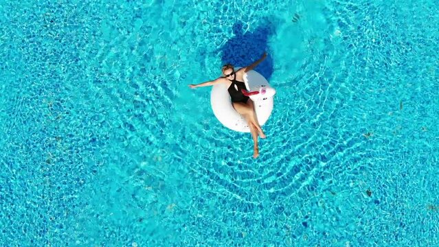  Woman relaxing in pool float unicorn inflatable ring floating on turquoise pool water. Aerial top view from drone.