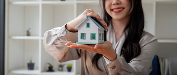 Smiling real estate agent woman hands protected miniature house on wooden desk. Property insurance...
