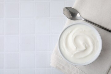 Delicious natural yogurt in bowl and spoon on white tiled table, top view. Space for text
