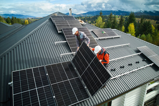 Roofers building photovoltaic solar module station on roof of house. Men electricians in helmets installing solar panel system outdoors. Concept of alternative and renewable energy. Aerial view.