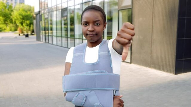 Young african american woman standing outside, broken arm clavicle fracture showing dislike thumb down gesture. Smiling beautiful girl patient near clinic hospital wear bandage arm brace relive pain
