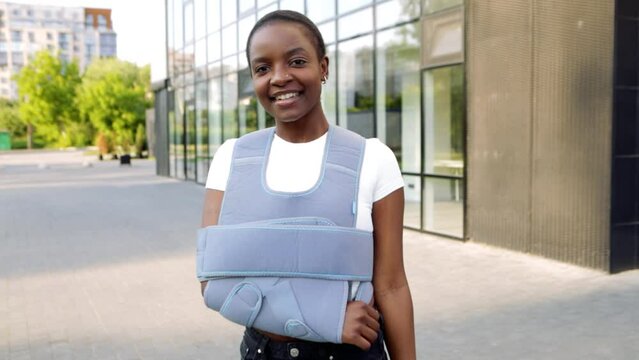 Young african american woman standing outside with broken arm clavicle fracture. Smiling beautiful girl patient near clinic hospital wear bandage arm brace to relive pain. Injury, accident, recovery