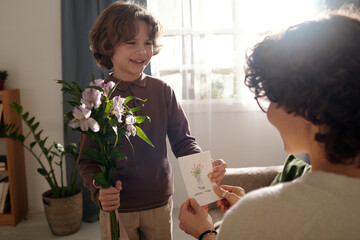Happy cute boy with bunch of flowers passing handmade greeting card to his mother while...
