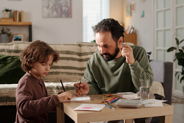 Young man with paintbrush in hand looking at his son drawing with crayons while both preparing handmade postcard for mother day