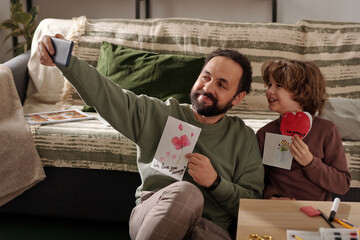 Happy young man and his son with handmade postcards looking at smartphone screen while taking...