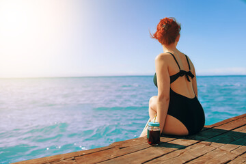 Attractive girl enjoys sea view on vacation. Red-haired woman in swimsuit is sitting on edge of a wooden pier with a bottle of refreshing lemonade next to her. Tourism and travel. Sunlight at sky.