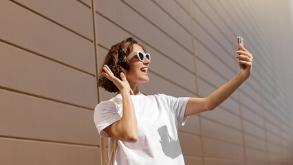 Happy young woman in stylish sunglasses taking selfie photo on smartphone on sunny summer day. Female blogger uses mobile phone camera, attractive caucasian girl touching hair for selfie picture.