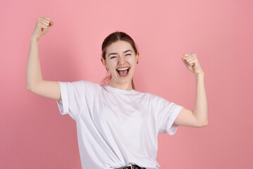 Attractive Caucasian young brunette woman in a casual white t-shirt screams with happiness and shows a winning gesture isolated on a pink studio background. - obrazy, fototapety, plakaty