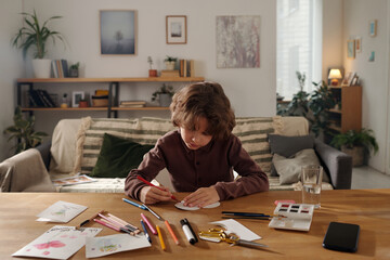 Diligent boy with red crayon creating handmade postcard for his mother while sitting by table with papers, watercolors and highlighters