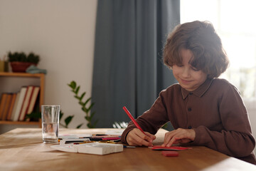 Little boy drawing on handmade paper postcard with crayons while sitting by table in front of camera and preparing gift for his mother