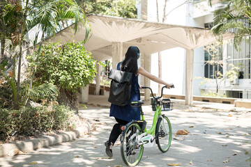 A young Asian woman commutes to work by cycling in a green city, carrying a backpack and using a...