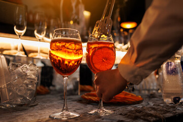Bartender makes two glasses of cocktail Aperol spritz on bar counter, adds fresh orange slices. Typical alcoholic Italian beverage, aperitif made with Prosecco sparkling white wine and ice cubes.