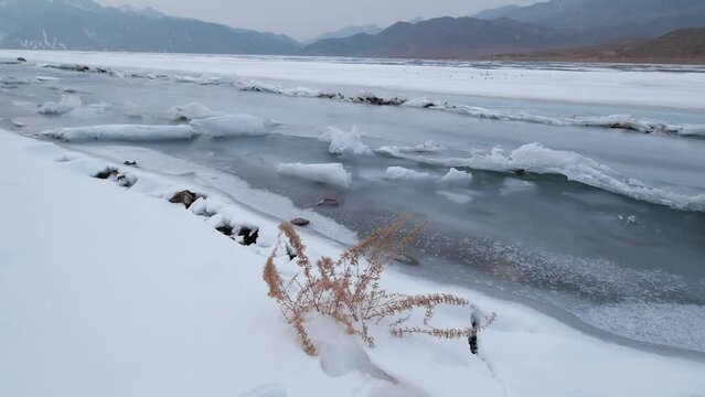 The wind blows dry grass onto a mountain lake covered with ice, snow and hummocks in early spring. Bartogay reservoir in Kazakhstan.