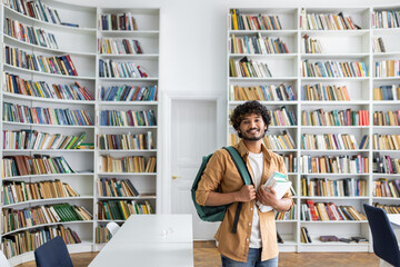 A cheerful young adult with curly hair and a backpack standing in a bright library, holding several...